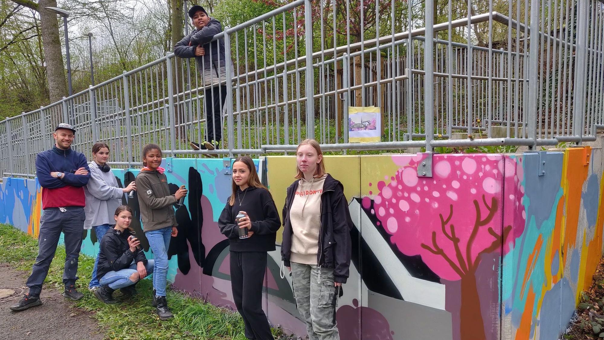 Die Graffiti-Künstler stehen mit dem ehrenamtlichen Workshop-Leiter Hannes Fries an der besprühten Mauer neben der Opladener Straße.