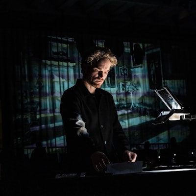 Ein Mann steht in einem dunklen Raum auf der Bühne und blickt nach unten. Er trägt schwarze Kleidung.