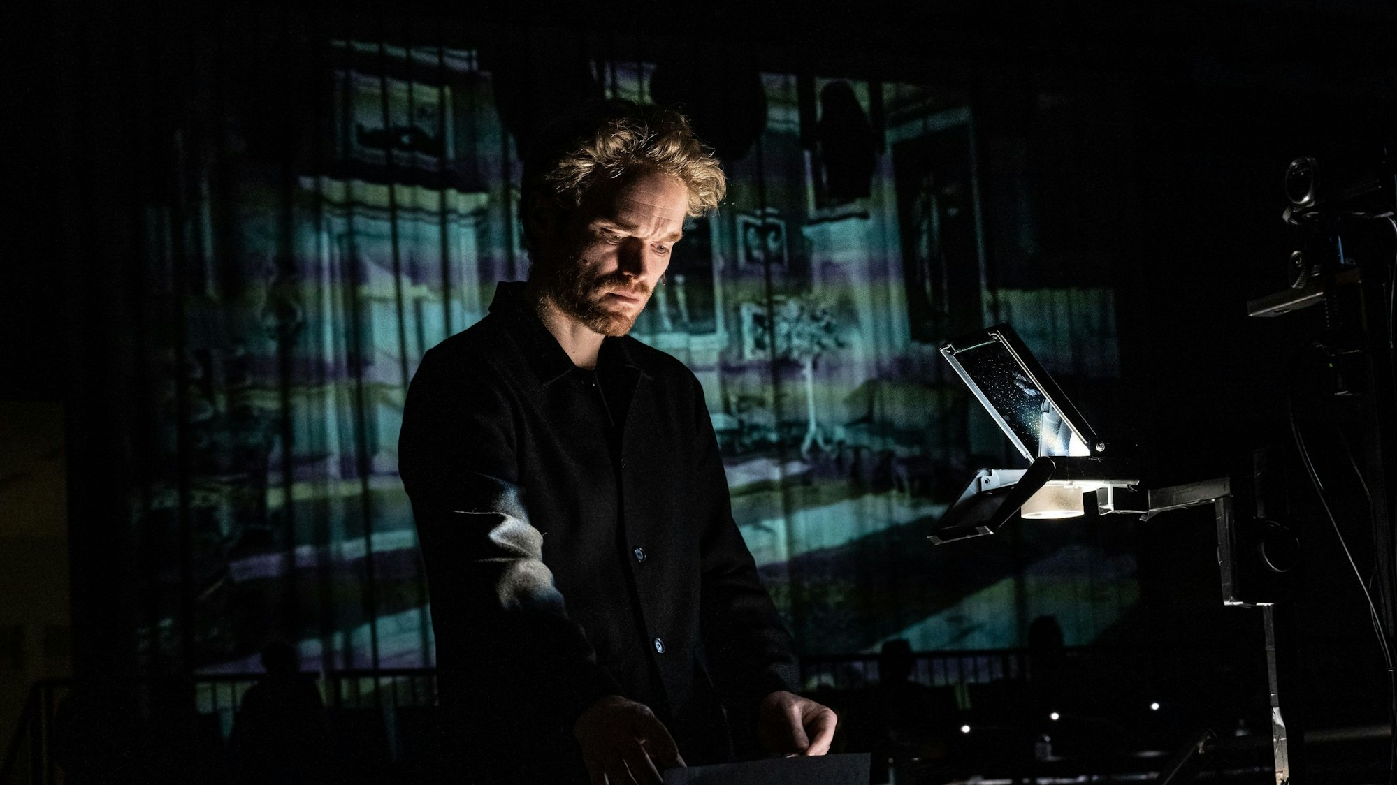 Ein Mann steht in einem dunklen Raum auf der Bühne und blickt nach unten. Er trägt schwarze Kleidung.