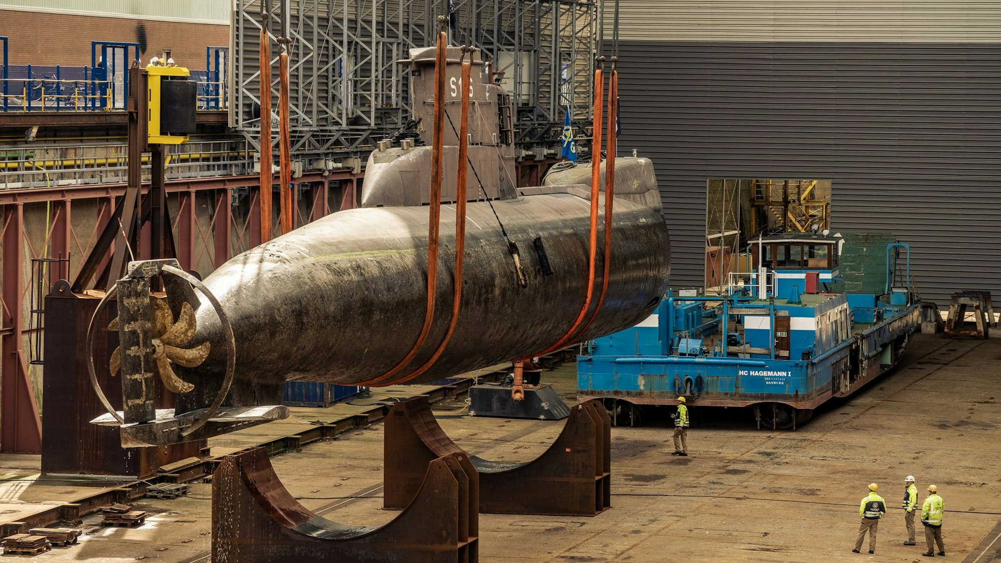 Das U-Boot in großen Trageschlaufen wird auf ein Trockendock gesetzt. Unten rechts beaufsichtigen drei Mitarbeiter den Transport.