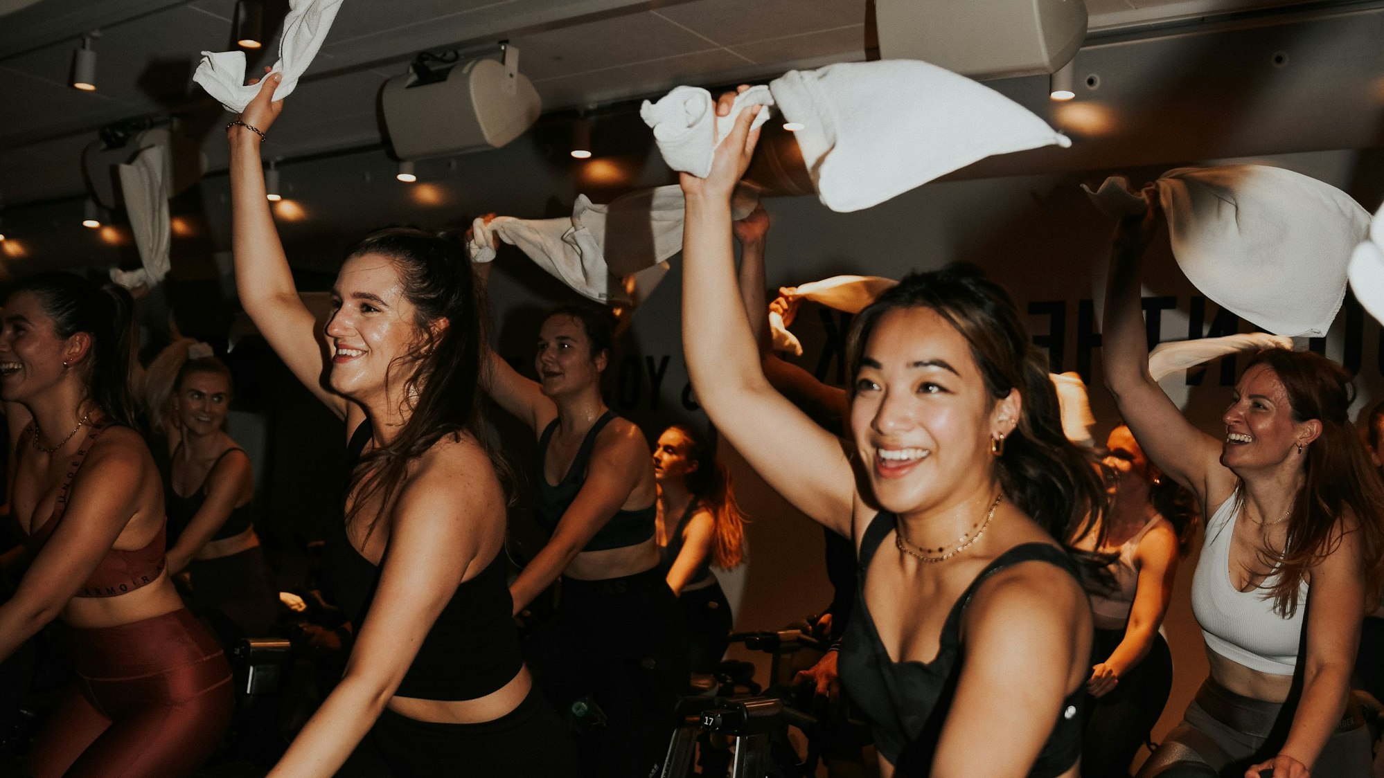 Frauen beim Spinning in einem dunklen Raum lachen und schwenken ihre Handtücher über dem Kopf.