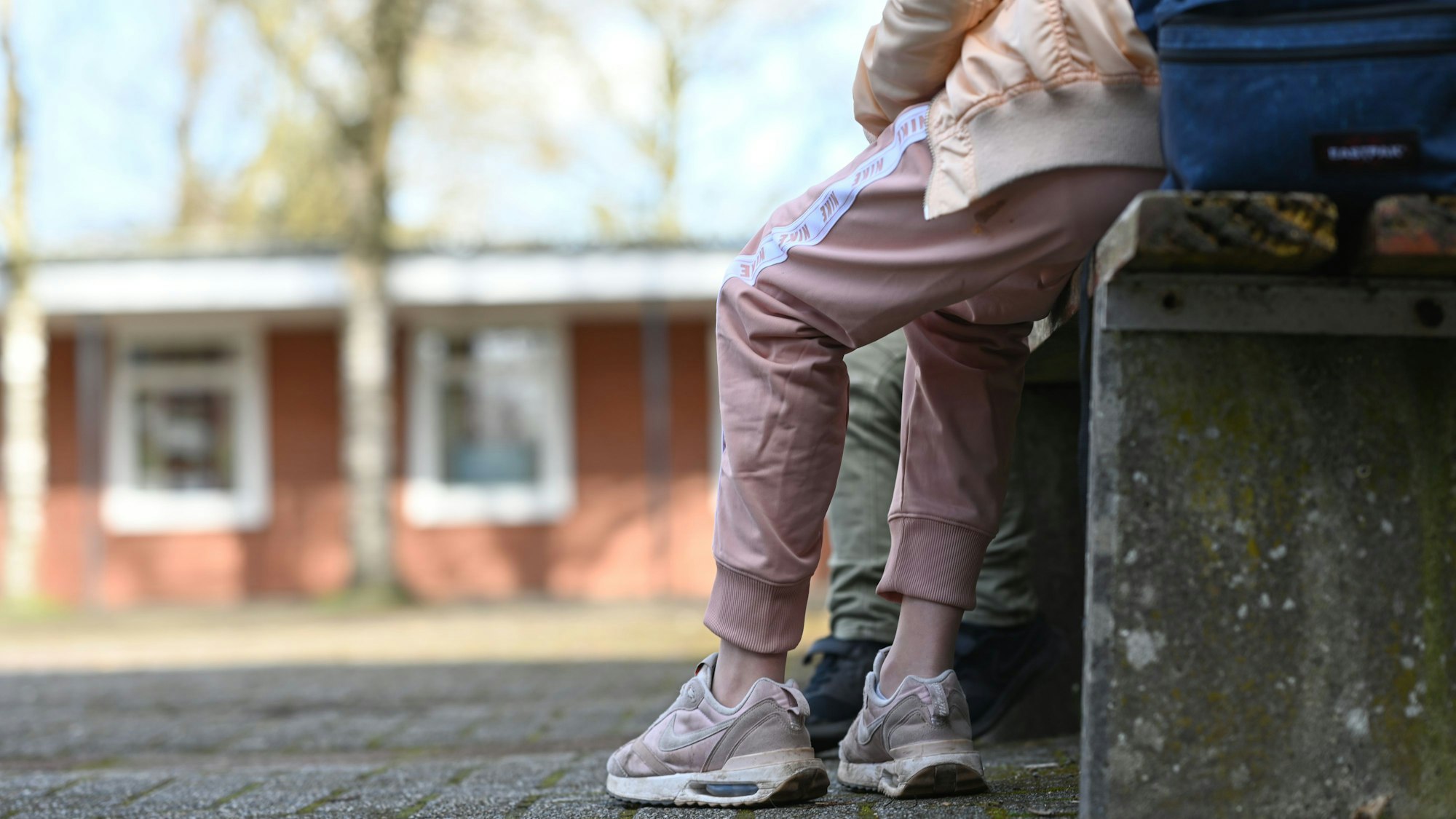 Eine Schülerin sitzt in einer rosafarbenen Jogginghose auf dem Schulhof einer Schule. Im Hintergrund ist ein einstöckiges Schulgebäude zu sehen.