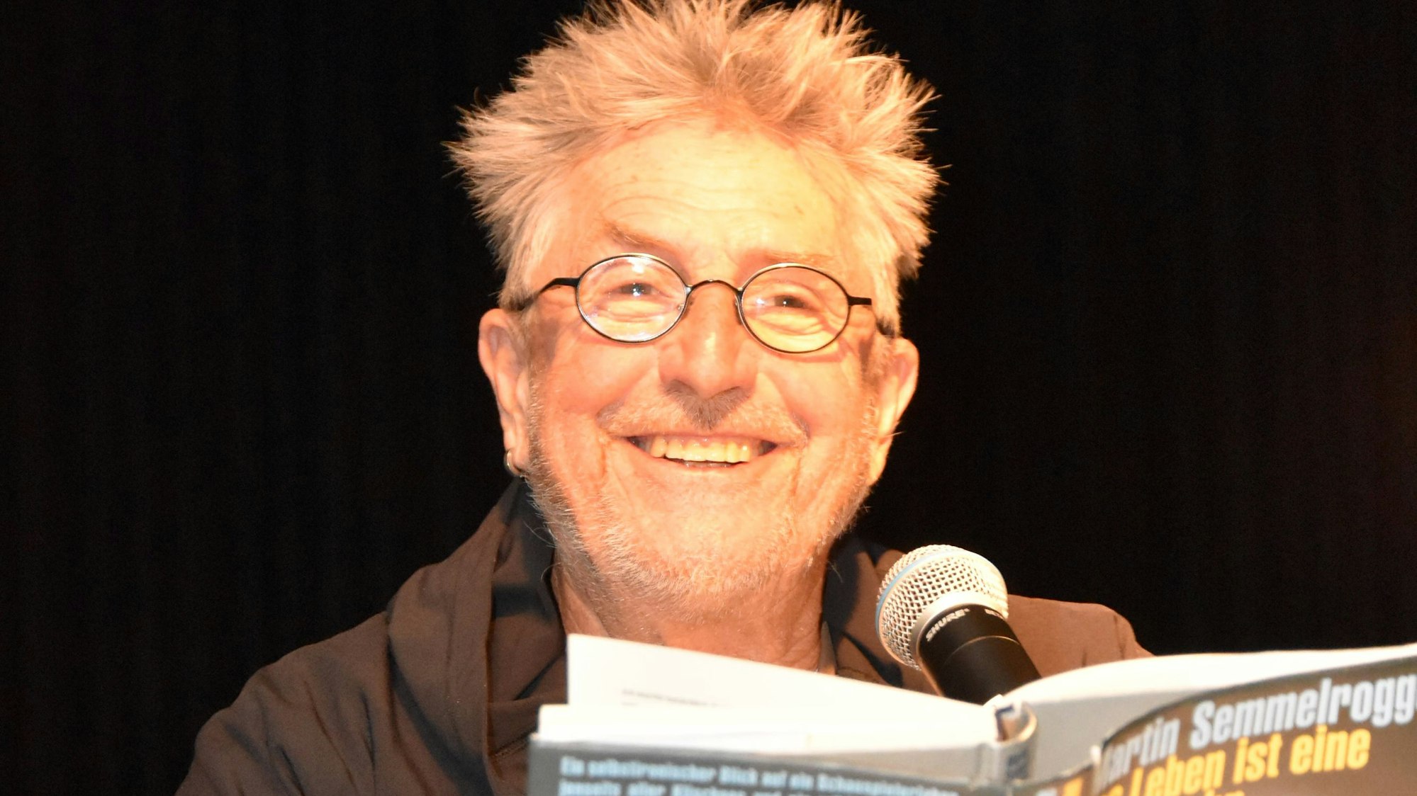 Semmelrogge sitzt an einem Tisch, hält seine Biografie in den Händen und lacht.