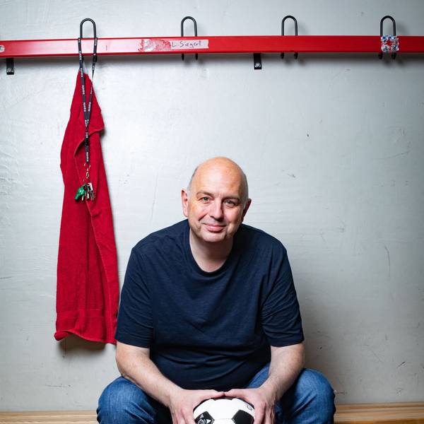 Frank Goosen sitzt auf einer Bank, in einem blauen T-Shirt vor einer weißen Wand. Links von ihm hängt ein rotes Handtuch und ein Schlüsselbund mit Trillerpfeife.