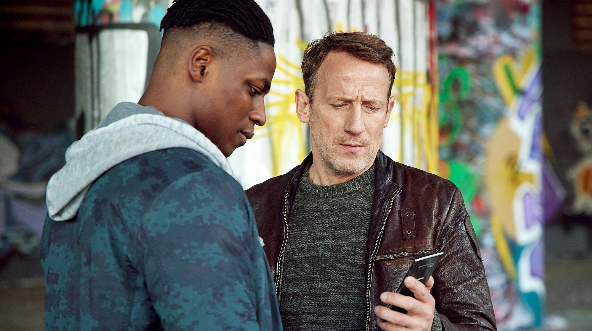 „Tatort“-Kommissar Thorsten Falke (Wotan Wilke Möhring) zeigt Sam ein Bild des Toten auf seinem Handy.