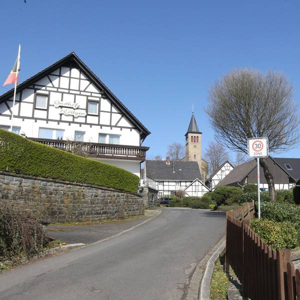 Ein Fachwerkhaus liegt an einer Straße, im Hintergrund ist eine Kirche zu sehen.