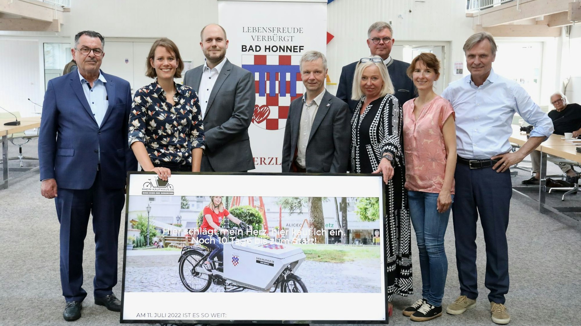 Acht Frauen und Männer stehen hinter einem großformatigen Monitor, auf dem eine Fahrerin mit einem Lastenrad zu sehen ist, mit dem das „Rhein-Kaufhaus“ Waren auslieferte.