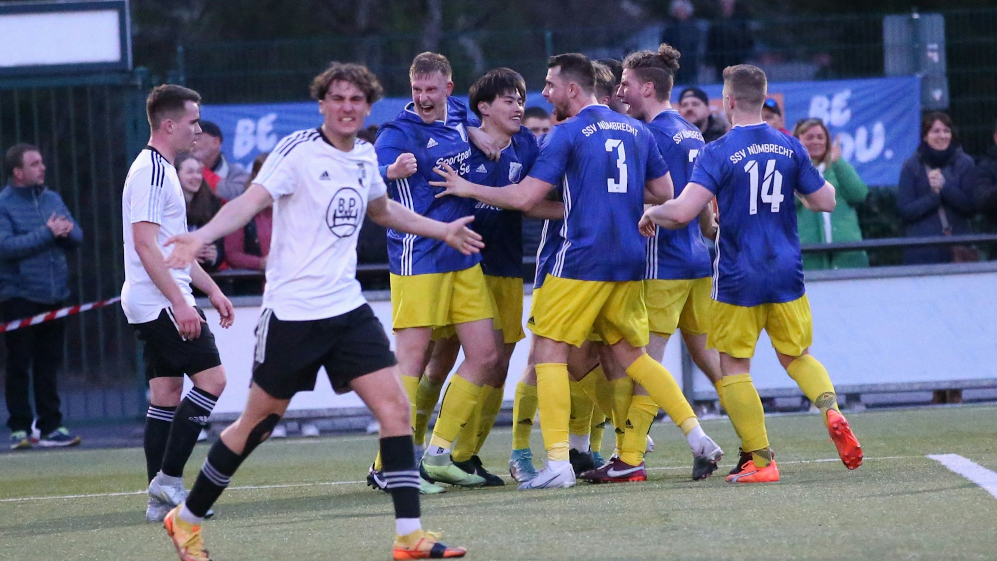 Bild einer jubelnden Fußball-Mannschaft, davor zwei skeptisch schauende Spieler der Gegenmannschaft.