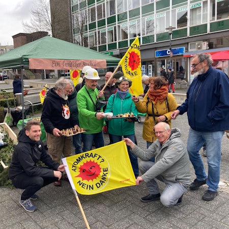Mehrere Personen stehen und knien, mit gelben Fahnen, auf denen „Atomkraft? Nein Danke!“ steht, in einer Fußgängerzone.