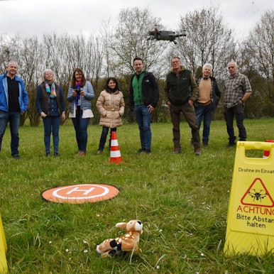 Mehrere Leute stehen auf einer Wiese in einer Reihe. Einer fliegt mit einer Drohne über ein im Gras liegendes Stoff-Rehkitz. Daneben stehen zwei gelbe Warnschilder mit der Aufschrift: „Drohne im Einsatz. Achtung, bitte Abstand halten“.