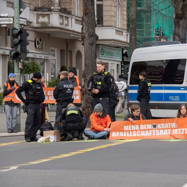 Aktivisten der Gruppe „Letzte Generation“ haben sich auf der Neuen Kantstraße auf einer Kreuzung festgeklebt. Die Polizei sichert den Bereich, während eine Polizistin begonnen hat, die Aktivisten von der Straße zu lösen.