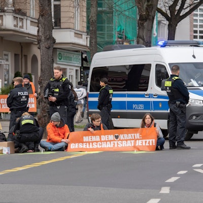 Aktivisten der Gruppe „Letzte Generation“ haben sich auf der Neuen Kantstraße auf einer Kreuzung festgeklebt. Die Polizei sichert den Bereich, während eine Polizistin begonnen hat, die Aktivisten von der Straße zu lösen.