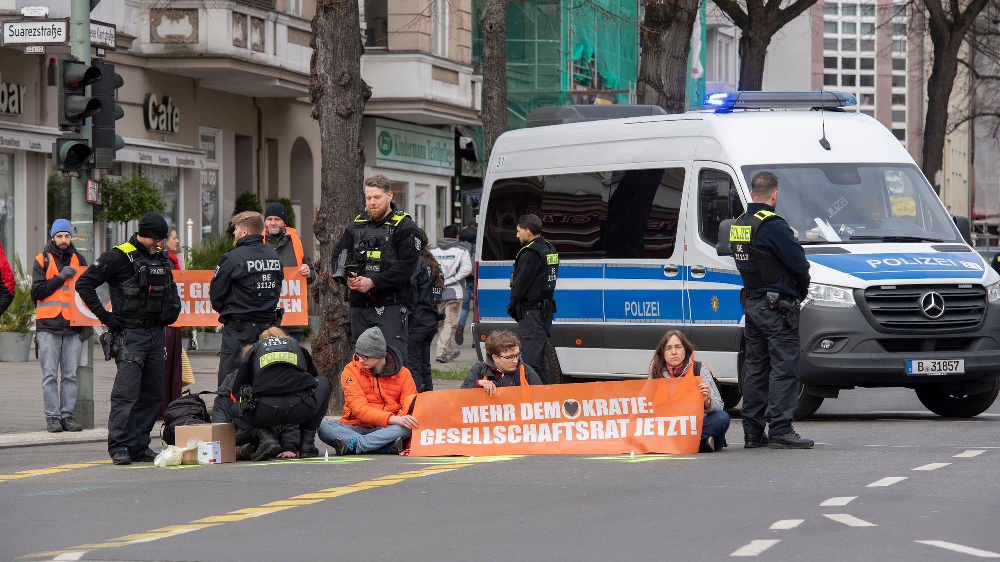 Aktivisten der Gruppe „Letzte Generation“ haben sich auf der Neuen Kantstraße auf einer Kreuzung festgeklebt. Die Polizei sichert den Bereich, während eine Polizistin begonnen hat, die Aktivisten von der Straße zu lösen.