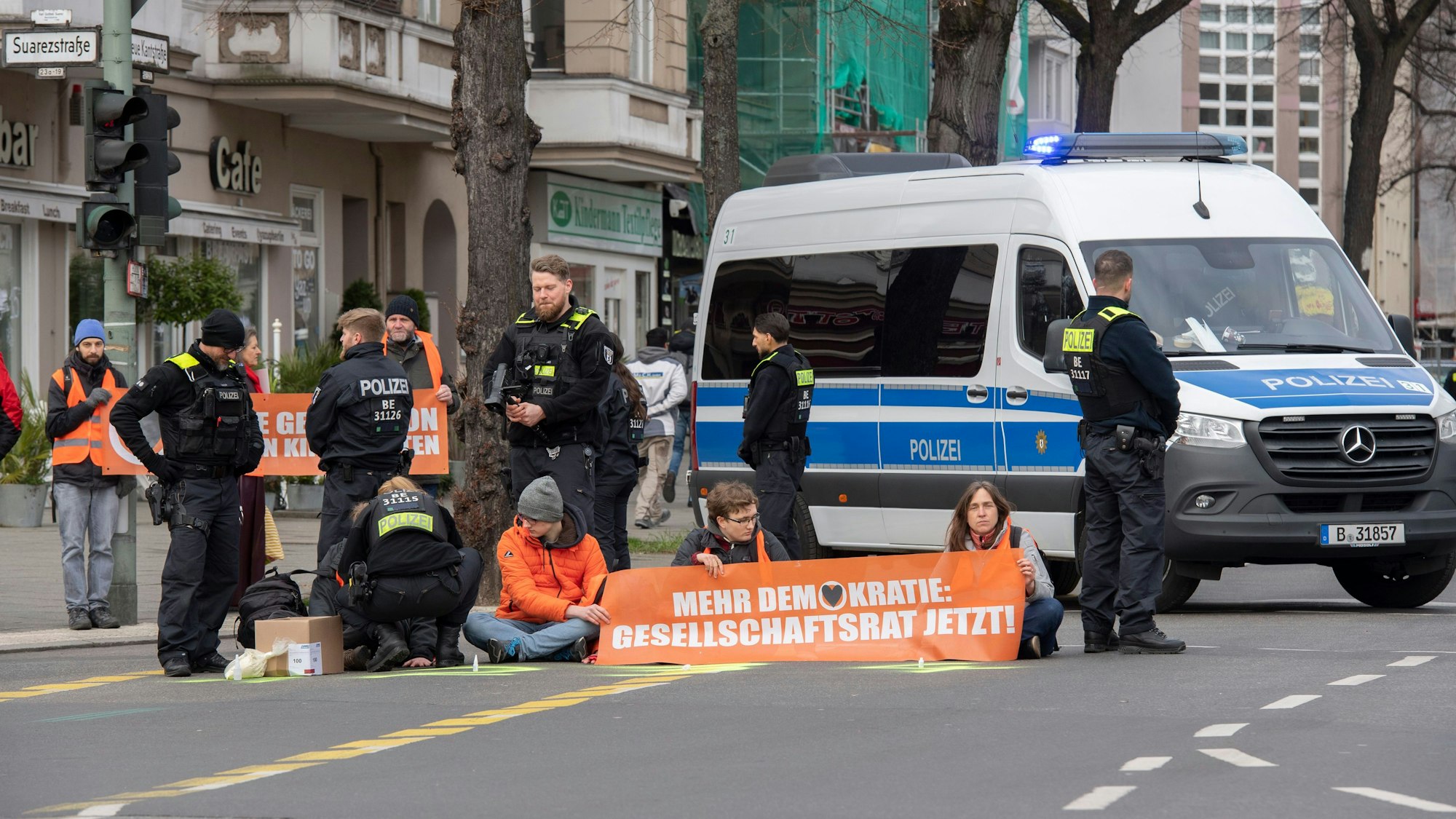 Aktivisten der Gruppe „Letzte Generation“ haben sich auf der Neuen Kantstraße auf einer Kreuzung festgeklebt. Die Polizei sichert den Bereich, während eine Polizistin begonnen hat, die Aktivisten von der Straße zu lösen.
