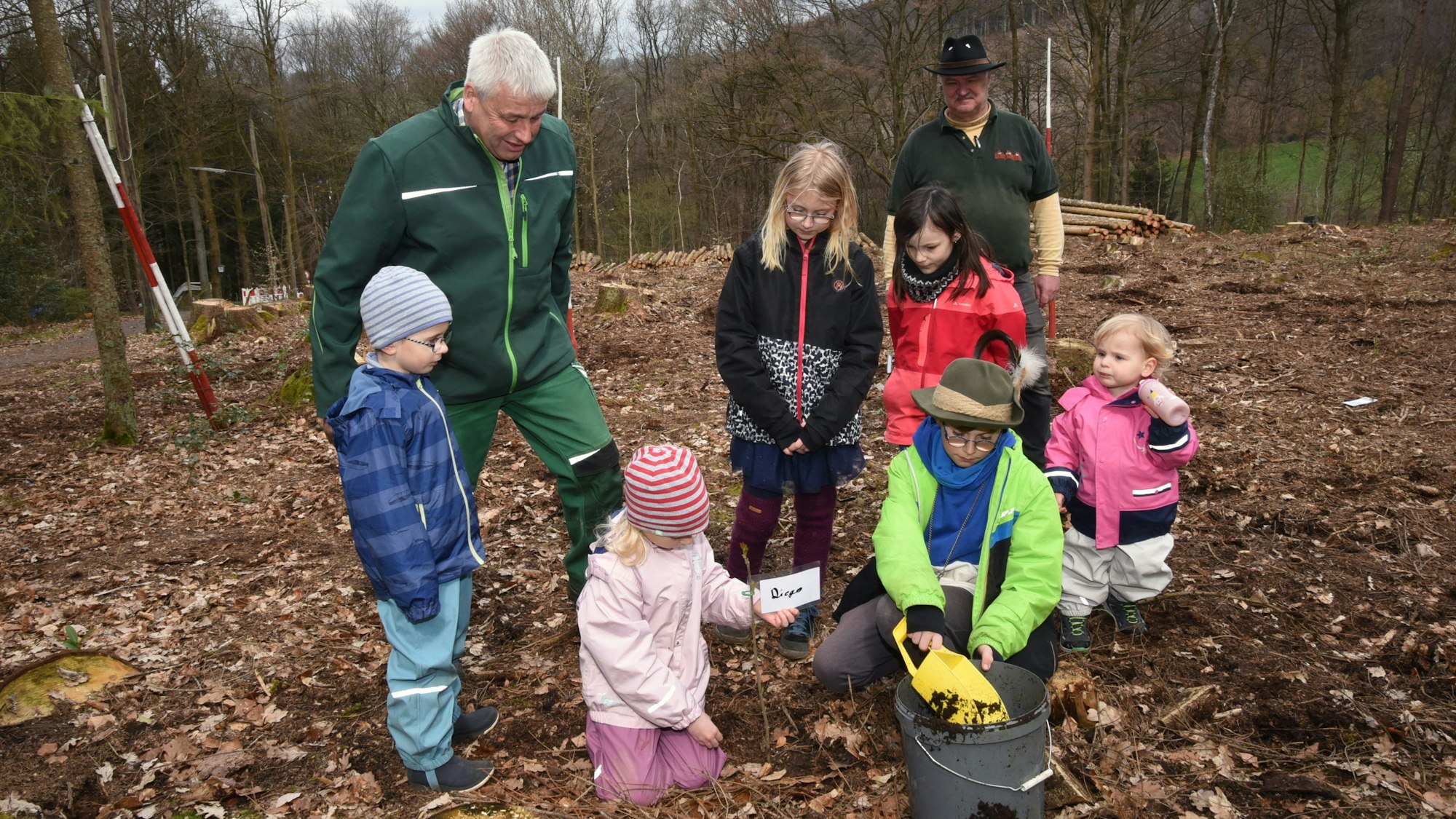 Mehrere Kinder pflanzen unter Anleitung eines älteren Mannes in grünem Overall kleine Setzlinge.