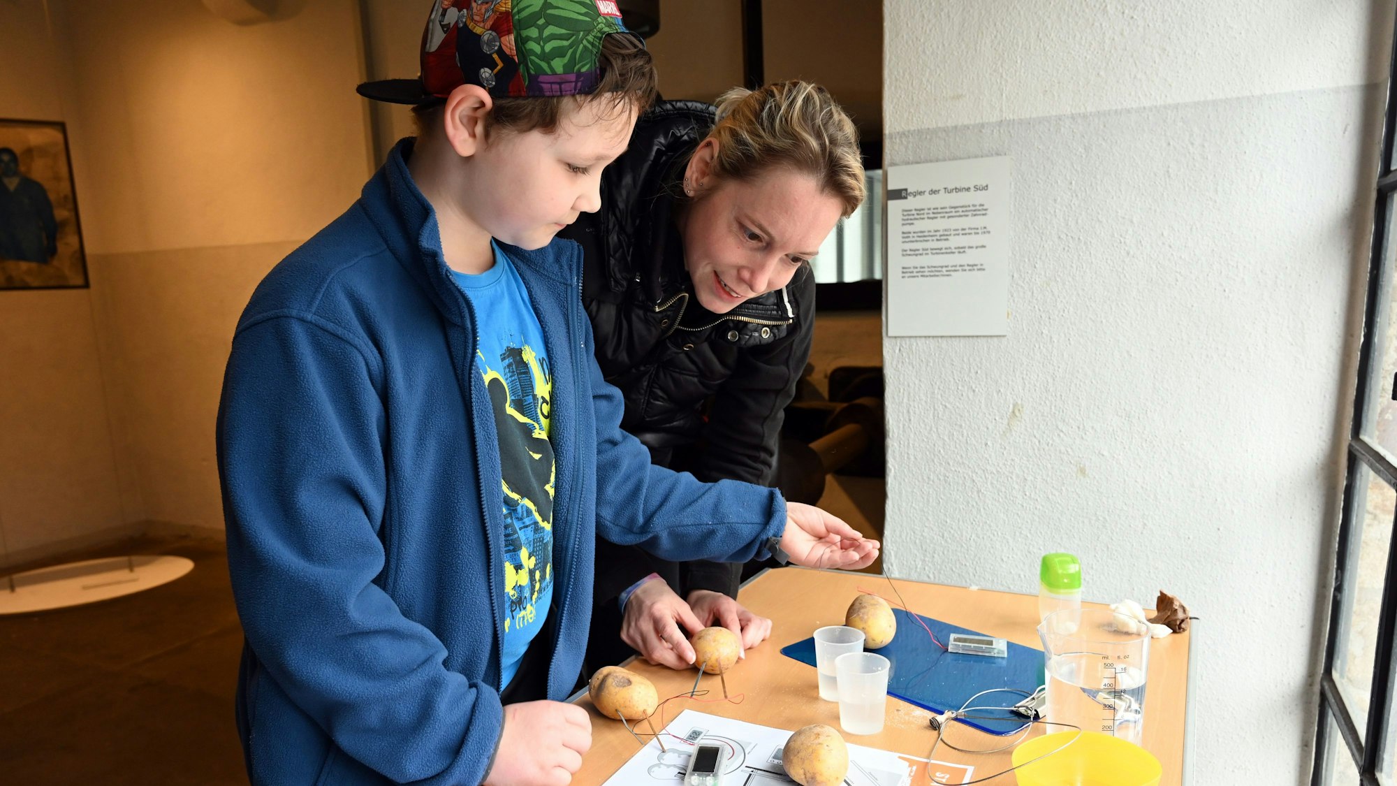 Ein Junge steckt Drähte und Kabel in eine rohe Kartoffel. Eine Frau schaut ihm dabei zu.