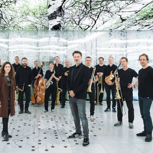 Heiner Schmitz und Veronika Morscher stehen etwas weiter vorne auf einem weißen Boden, auf dem ein stilisierter Baum aus Punkten und Linien abgebildet ist. Hinter ihnen steht ein großes Ensemble, alle sind in schwarz gekleidet. Über ihnen sind Bilder von Bäumen an der Decke.