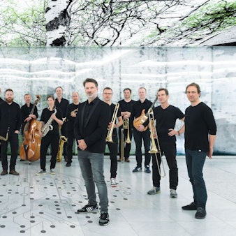 Heiner Schmitz und Veronika Morscher stehen etwas weiter vorne auf einem weißen Boden, auf dem ein stilisierter Baum aus Punkten und Linien abgebildet ist. Hinter ihnen steht ein großes Ensemble, alle sind in schwarz gekleidet. Über ihnen sind Bilder von Bäumen an der Decke.