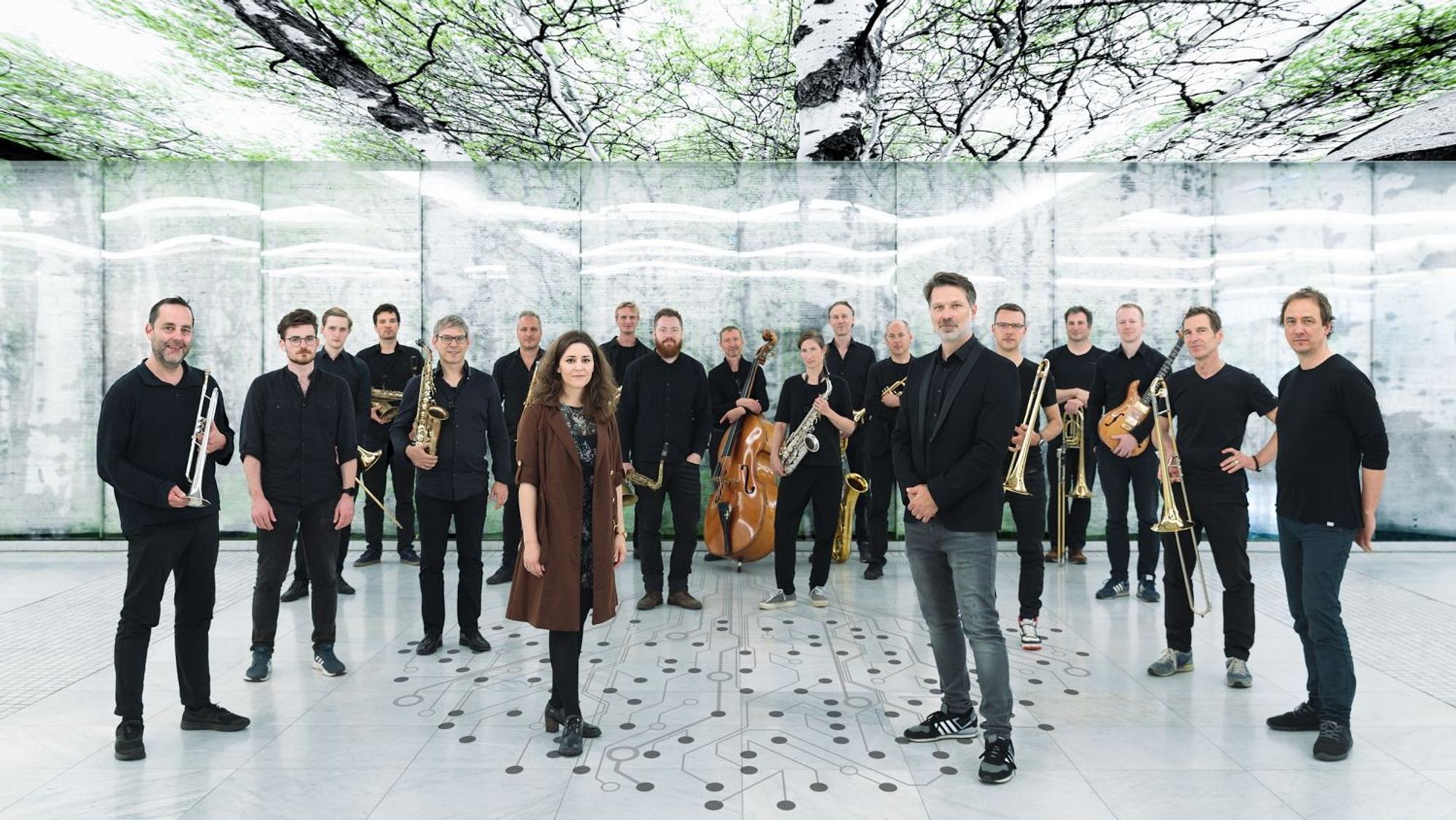 Heiner Schmitz und Veronika Morscher stehen etwas weiter vorne auf einem weißen Boden, auf dem ein stilisierter Baum aus Punkten und Linien abgebildet ist. Hinter ihnen steht ein großes Ensemble, alle sind in schwarz gekleidet. Über ihnen sind Bilder von Bäumen an der Decke.