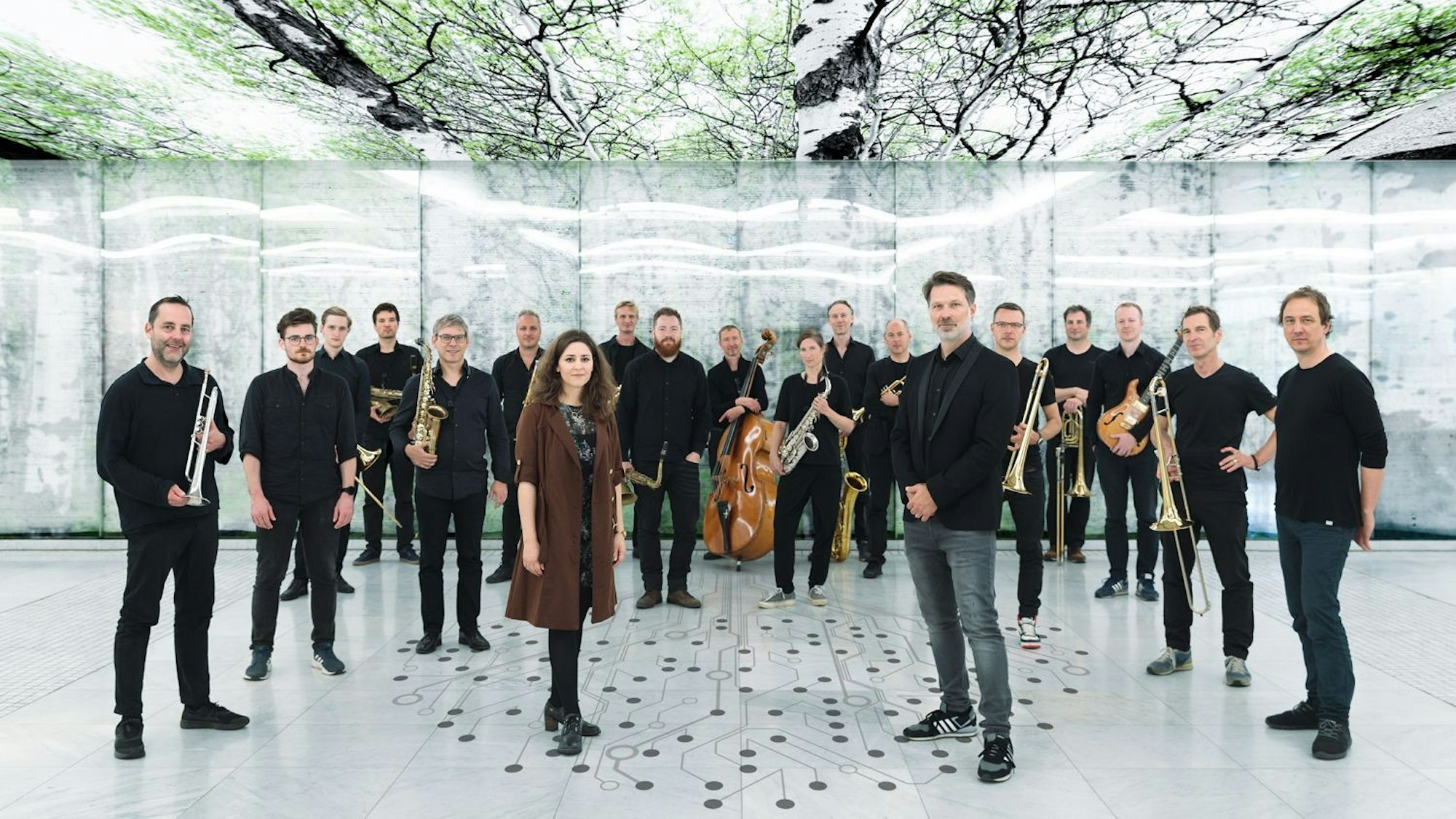 Heiner Schmitz und Veronika Morscher stehen etwas weiter vorne auf einem weißen Boden, auf dem ein stilisierter Baum aus Punkten und Linien abgebildet ist. Hinter ihnen steht ein großes Ensemble, alle sind in schwarz gekleidet. Über ihnen sind Bilder von Bäumen an der Decke.