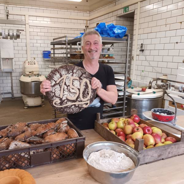 Es ist „Fibbes“ zu sehen, der ein großes Brot mit der Zahl 150 in die Kamera hält.