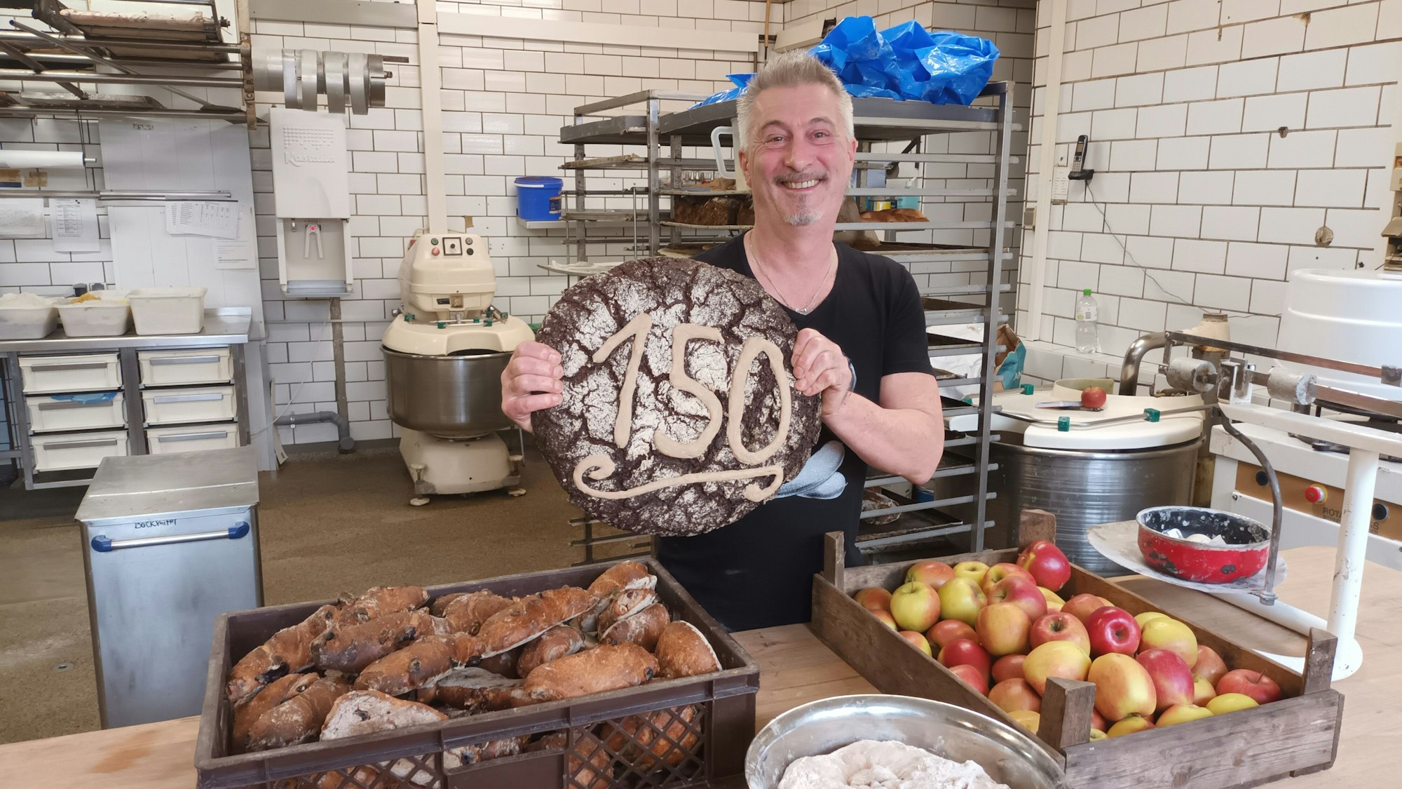 Es ist „Fibbes“ zu sehen, der ein großes Brot mit der Zahl 150 in die Kamera hält.