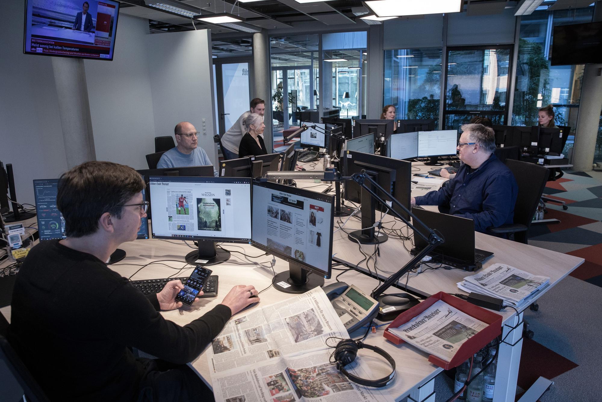 Redakteurinnen und Redakteure sitzen an sternförmig angeordneten Schreibtischen und arbeiten an PC mit mehreren Bildschirmen.