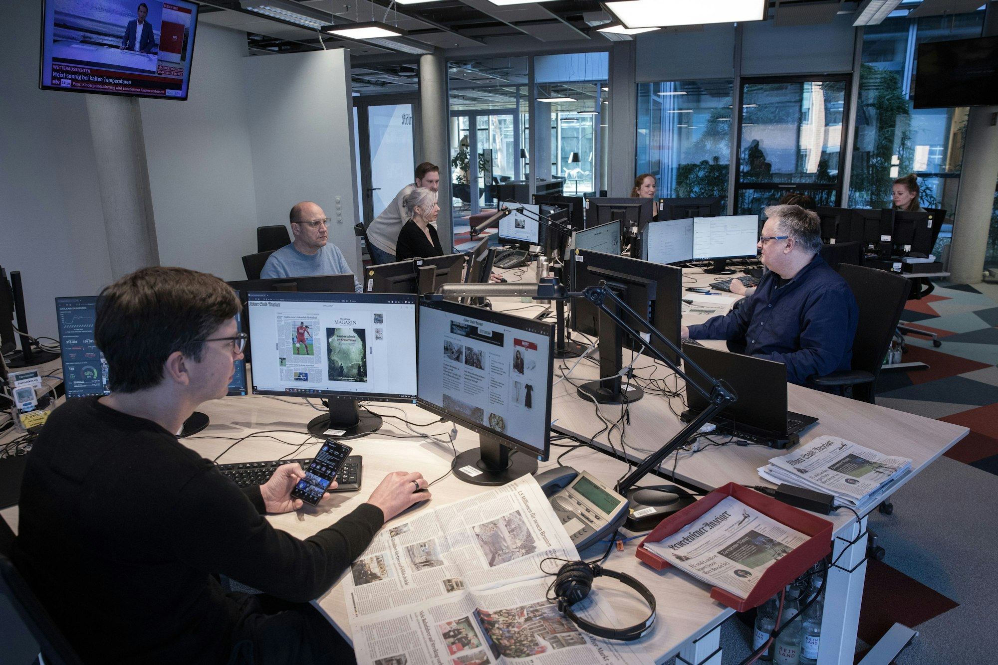 Redakteurinnen und Redakteure sitzen an sternförmig angeordneten Schreibtischen und arbeiten an PC mit mehreren Bildschirmen.