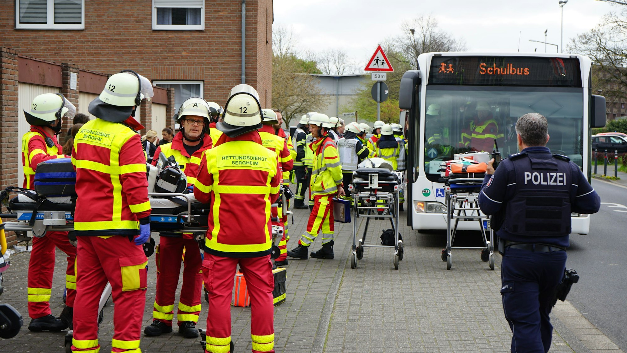 Rettungskräfte und ein Polizist stehen vor einem Gebäude. Ein Schulbus steht am Straßenrand.