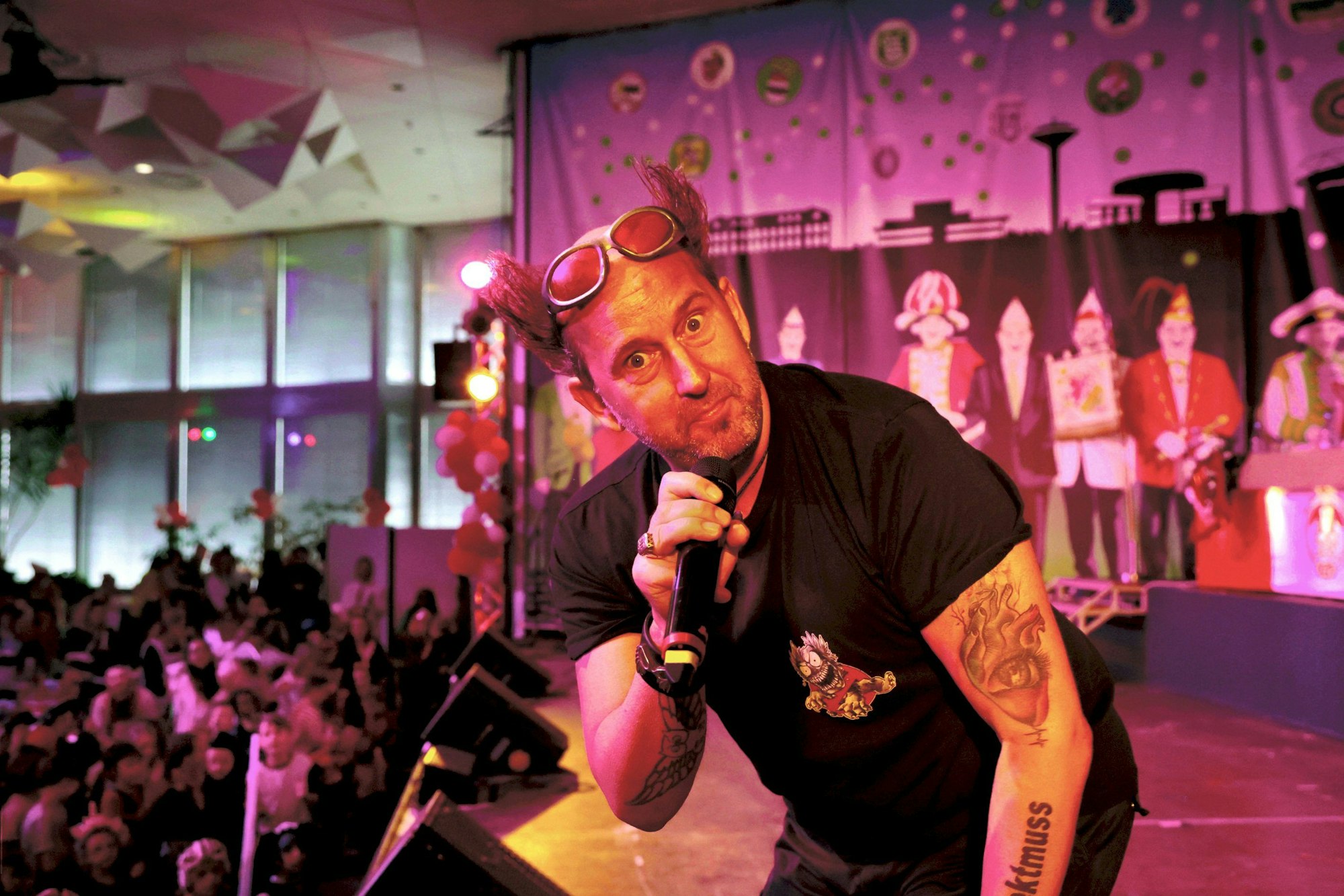 KIKA-TV Star Tom Lehel steht auf einer pink beleuchteten Bühne. Er trägt ein schwarzes T-Shirt und eine hochgeschobene Sonnenbrille. In der Hand hält er ein Mikrofon, er schaut spaßhaft in die Kamera.
