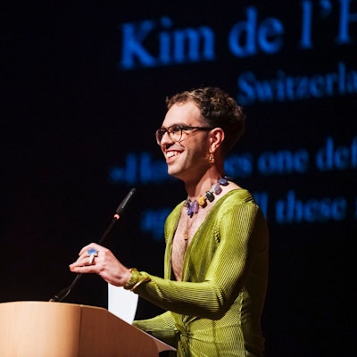 Kim de l'Horizon steht am Mikrofon mit einem grünen Kleid und Schmuck.