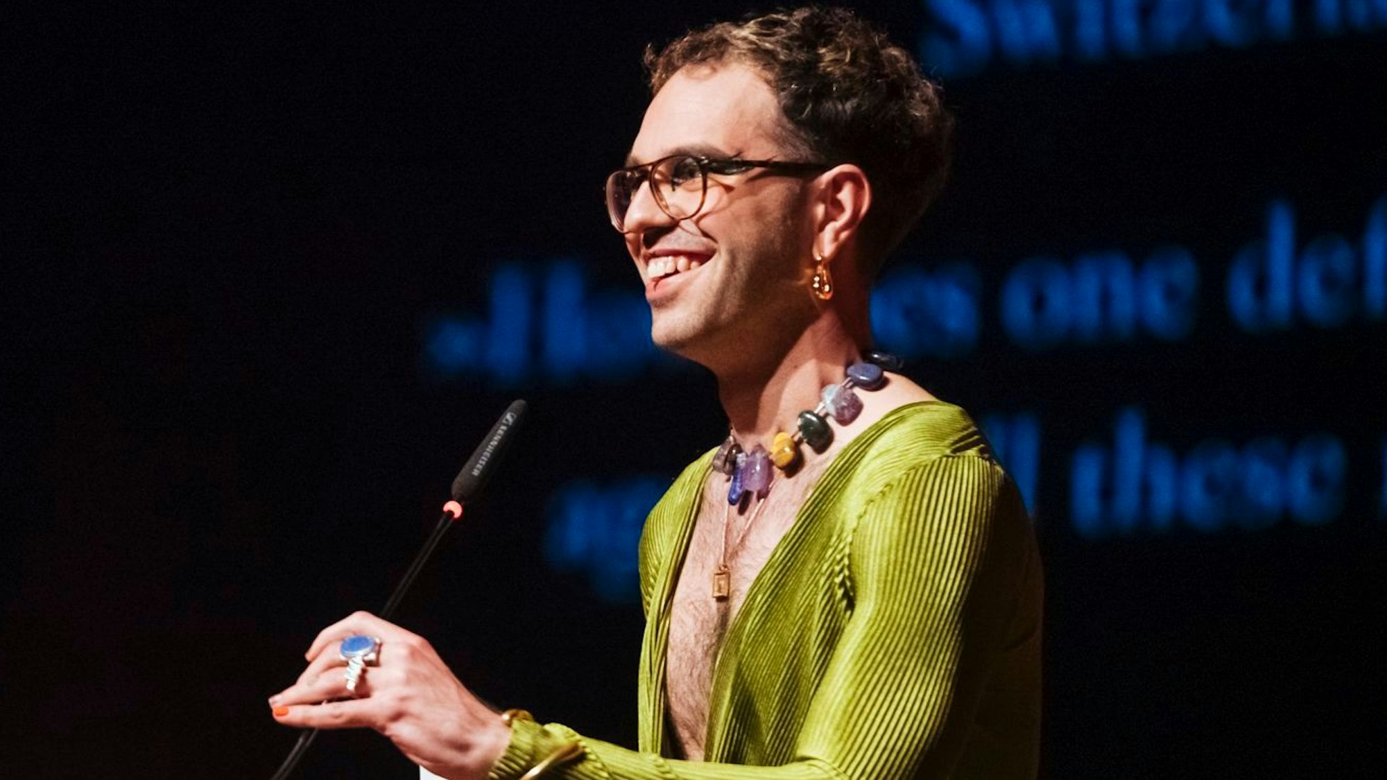 Kim de l'Horizon steht am Mikrofon mit einem grünen Kleid und Schmuck.