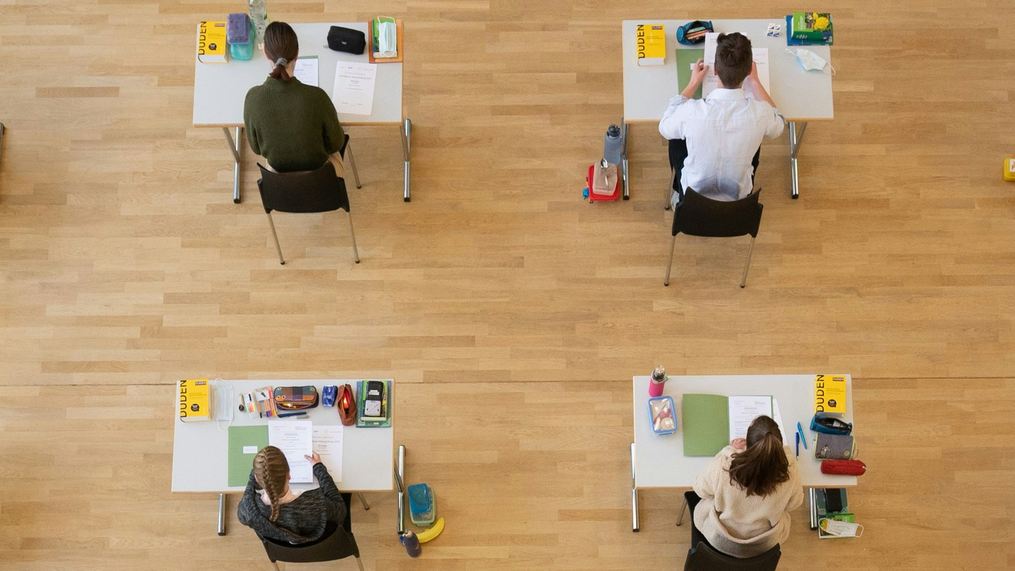 Schüler sind während der Abiturprüfung aus der Vogelperspektive zu sehen. Sie sitzen an weißen Tischen, auf denen je ein Duden liegt, Schreibzeug und die Aufgabenmappe.