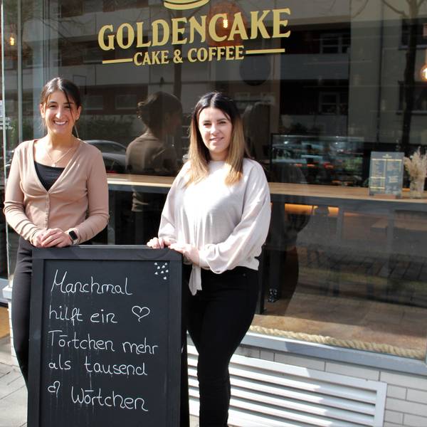 Zwei Frauen stehen vor der Glasfront eines Cafés. Sie stehen hinter einer Tafel, auf der steht Manchmal hilft ein Törtchen mehr als tausend Wörtchen.