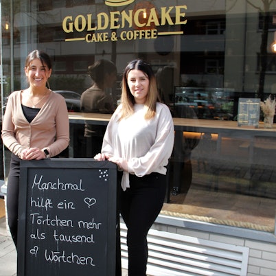 Zwei Frauen stehen vor der Glasfront eines Cafés. Sie stehen hinter einer Tafel, auf der steht Manchmal hilft ein Törtchen mehr als tausend Wörtchen.