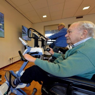 Eine Person im Vordergrund sitzt auf einem Sportgerät, während vor ihm ein Bildschirm steht. Im hinteren Teil des Raumes ist eine weitere Person auf einem anderen Sportgerät. Auch hier steht ein Bildschirm.