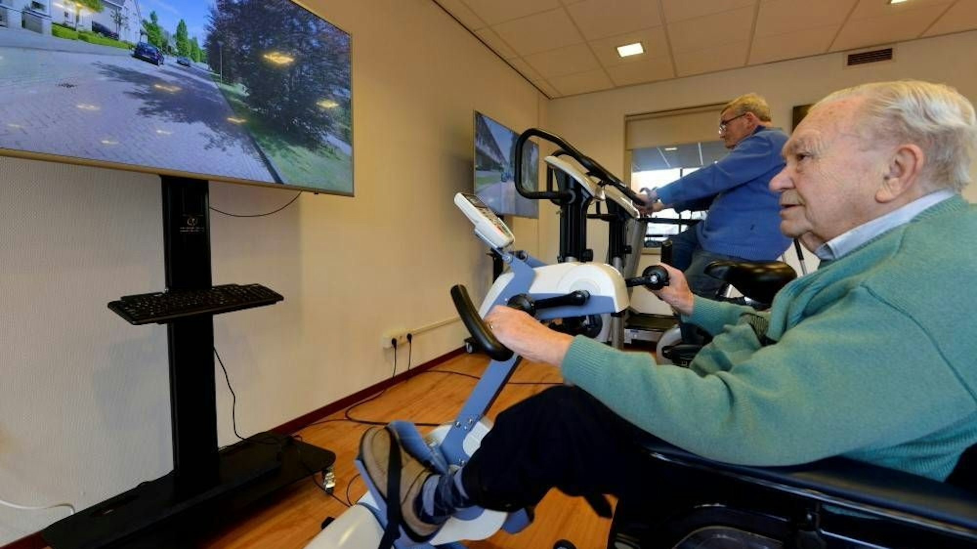 Eine Person im Vordergrund sitzt auf einem Sportgerät, während vor ihm ein Bildschirm steht. Im hinteren Teil des Raumes ist eine weitere Person auf einem anderen Sportgerät. Auch hier steht ein Bildschirm.