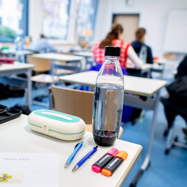 Ein Klassenraum mit mehreren Schülerinnen und Schülern, im Vordergrund ein Schreibtisch mit einem Bogen, auf dem „Abitur 2023“ steht.