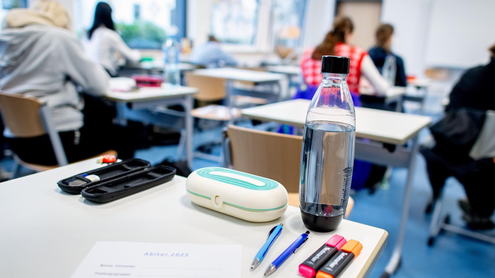 Ein Klassenraum mit mehreren Schülerinnen und Schülern, im Vordergrund ein Schreibtisch mit einem Bogen, auf dem „Abitur 2023“ steht.