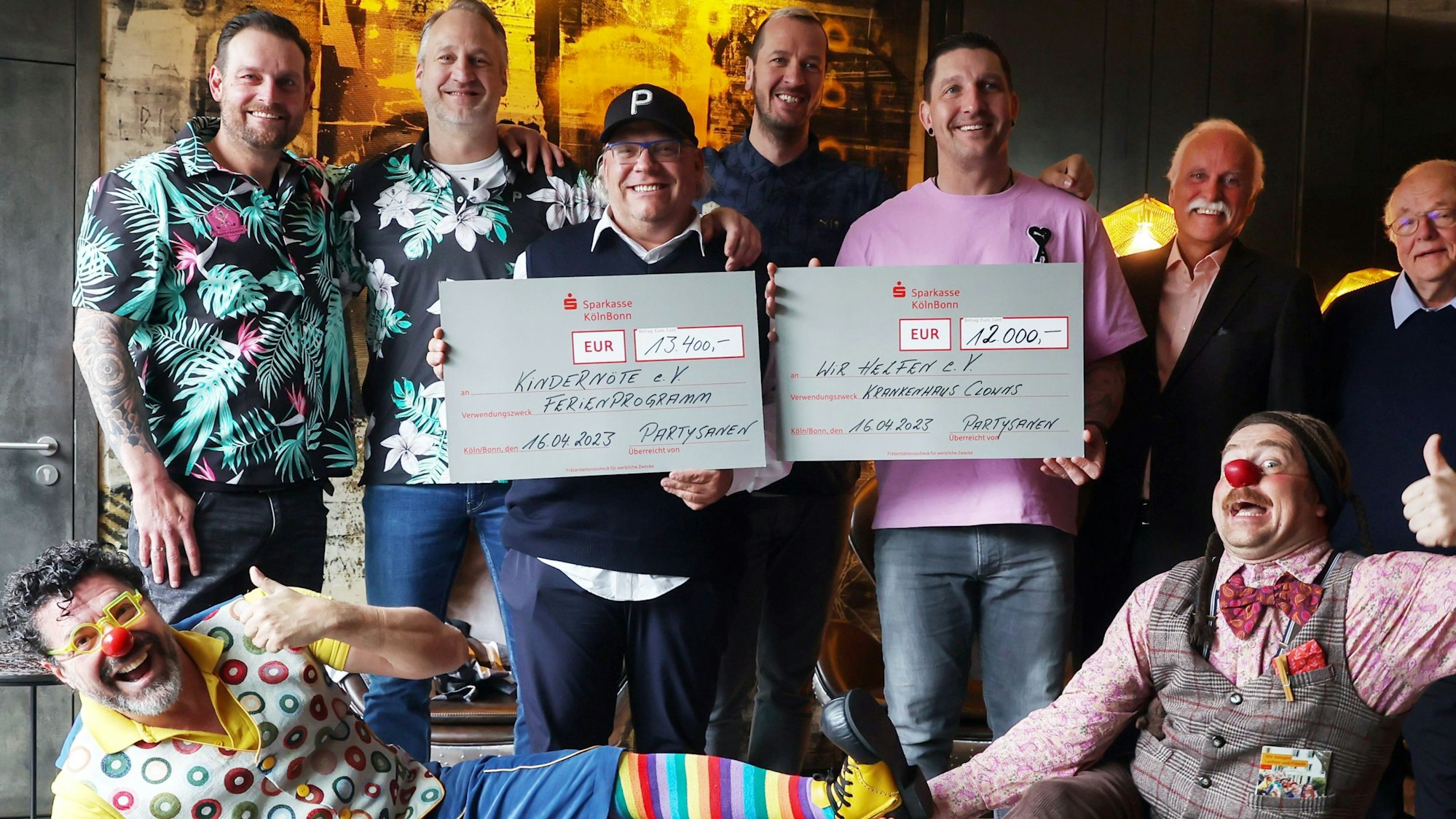 Die Sportlegenden Stefan Blank, Christian Ramota, Pascal Hens und Stefan Kretzschmar überreichen einen symbolischen Scheck in Höhe von 12.000 Euro an wir helfen und 13.400 Euro an den Kölner Verein Kindernöte e.V. Mit im Bild sind die beiden Klinikclowns Hallihallo und Lupo.