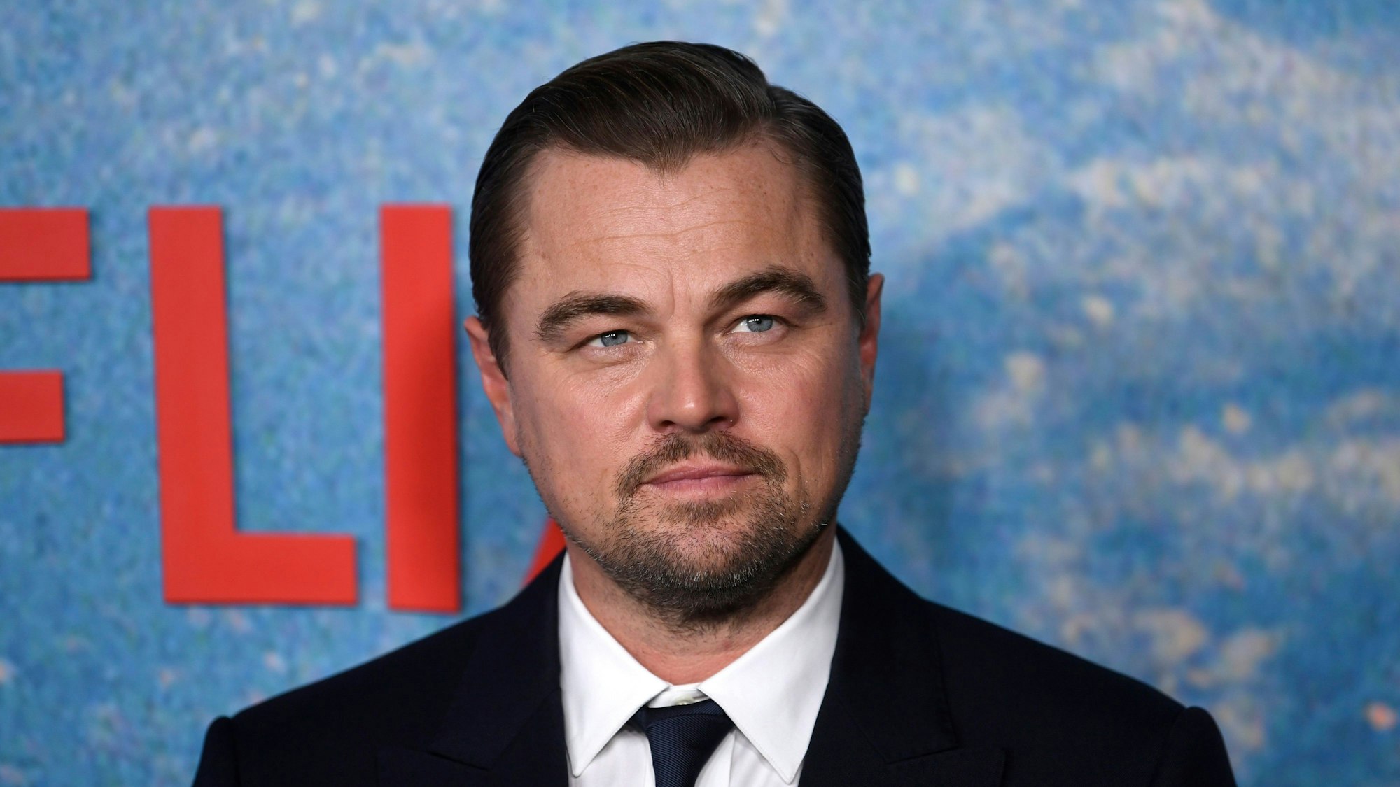 Schauspieler Leonardo Di Caprio blickt auf dem Roten Teppich in die Kamera. Er trägt einen schwarzen Anzug.