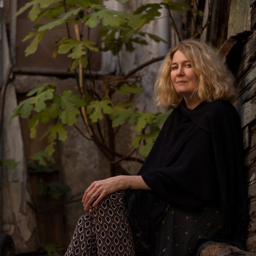 Sabine Schiffner sitzt vor einer grauen, von einer Pflanze berankten, Außenwand. Sie trägt ein schwarzes Oberteil und blickt in die Kamera