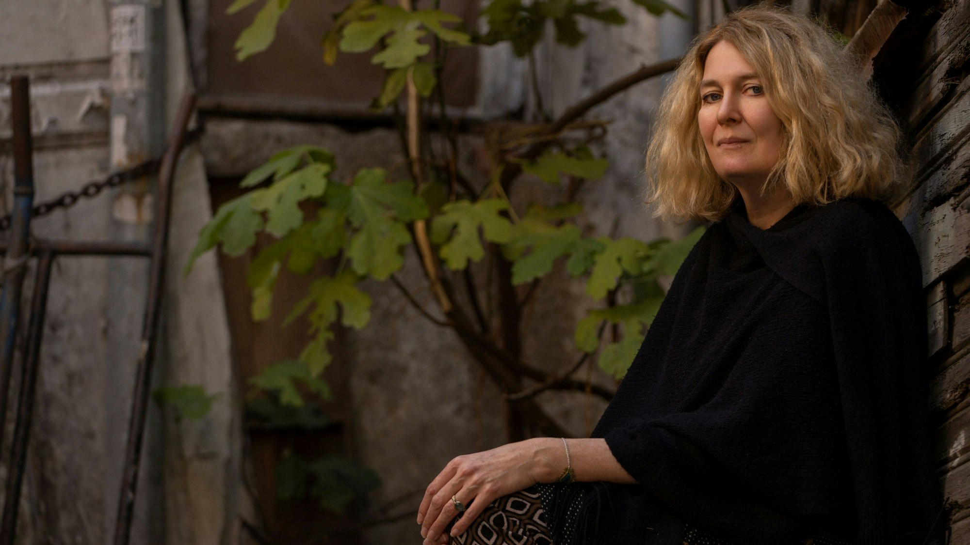 Sabine Schiffner sitzt vor einer grauen, von einer Pflanze berankten, Außenwand. Sie trägt ein schwarzes Oberteil und blickt in die Kamera