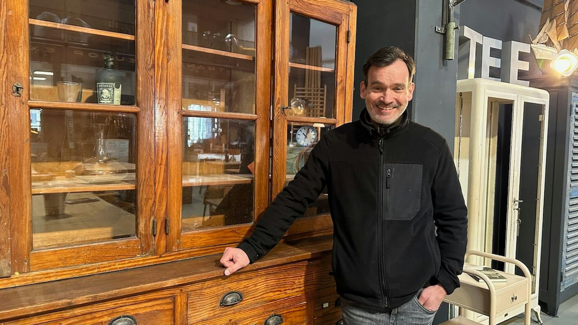 Mann vor einer antik aussehenden Kommode mit Glasvitrine