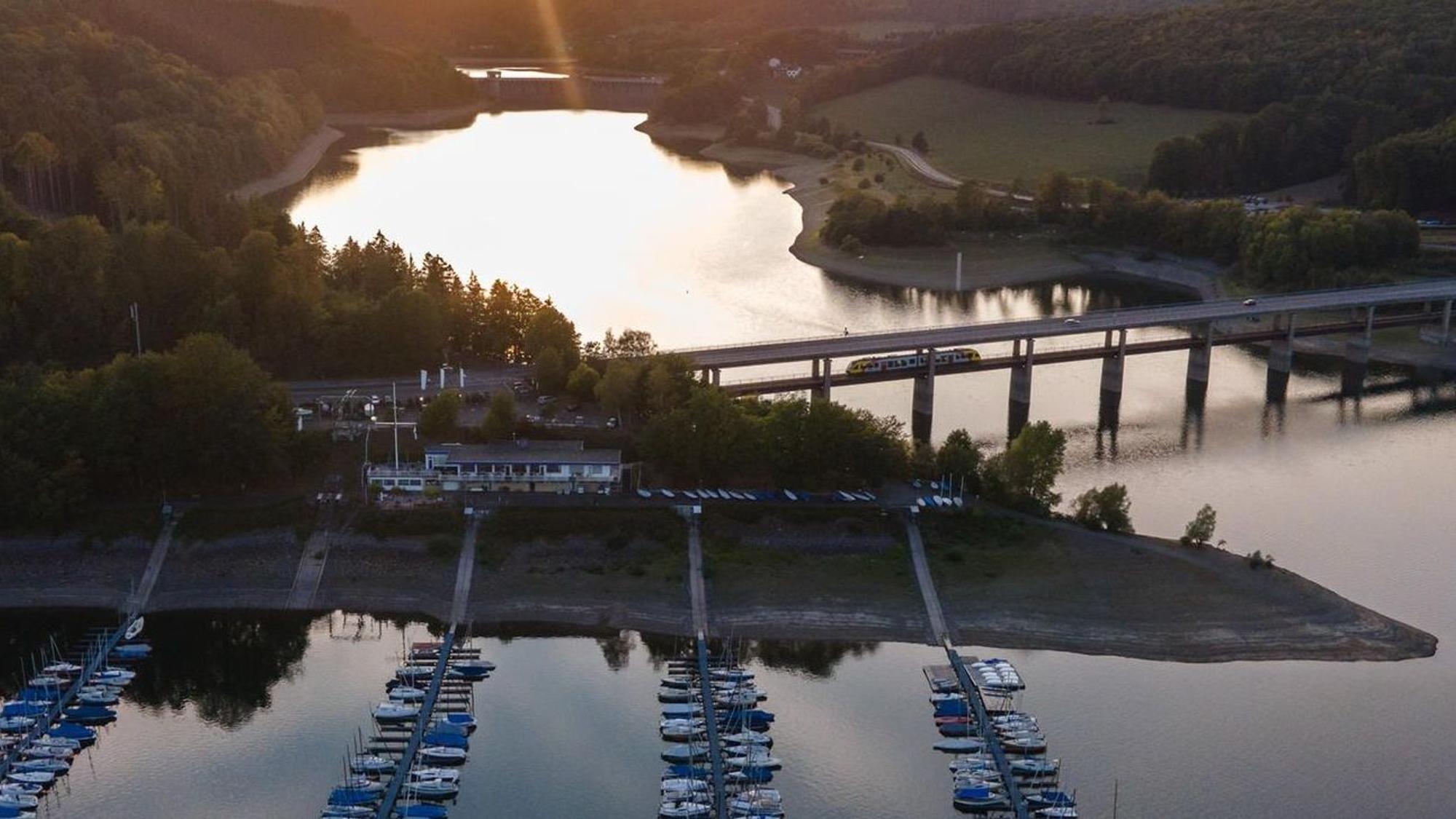 Die Sonne geht am Biggesee hinter einem Segelhafen und einer Brücke unter.
