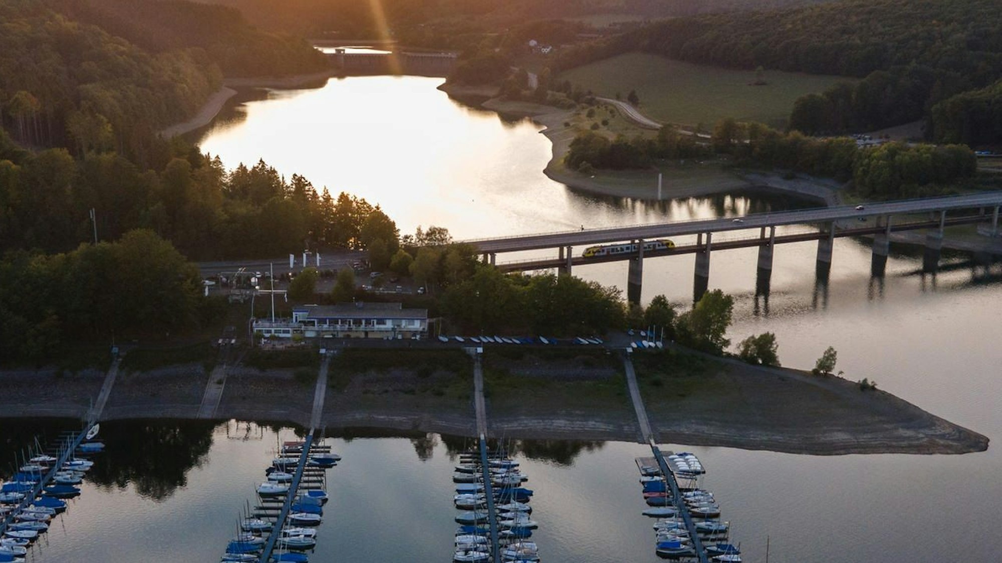Die Sonne geht am Biggesee hinter einem Segelhafen und einer Brücke unter.