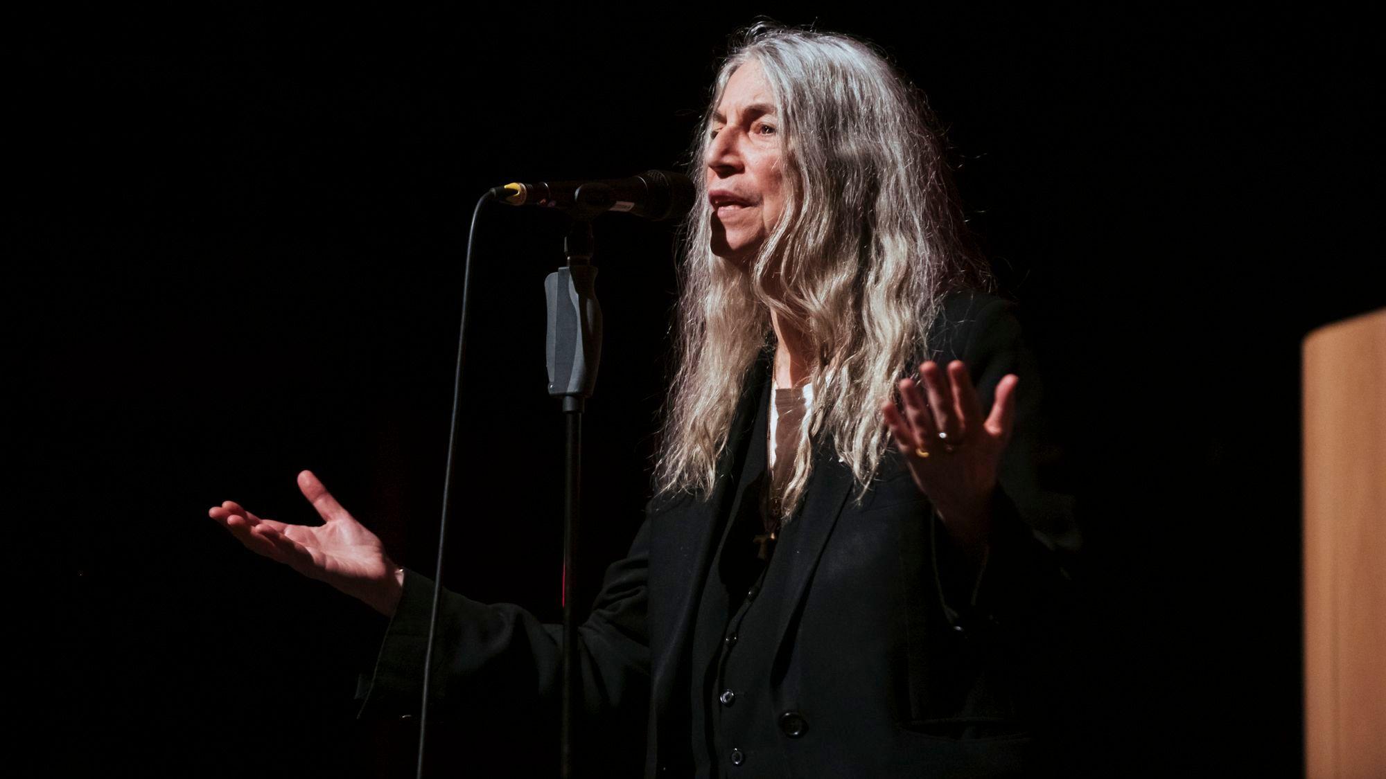 Patti Smith steht mit ausgestreckten Händen vor einem Mikrofonständer und trägt ein Gedicht vor.