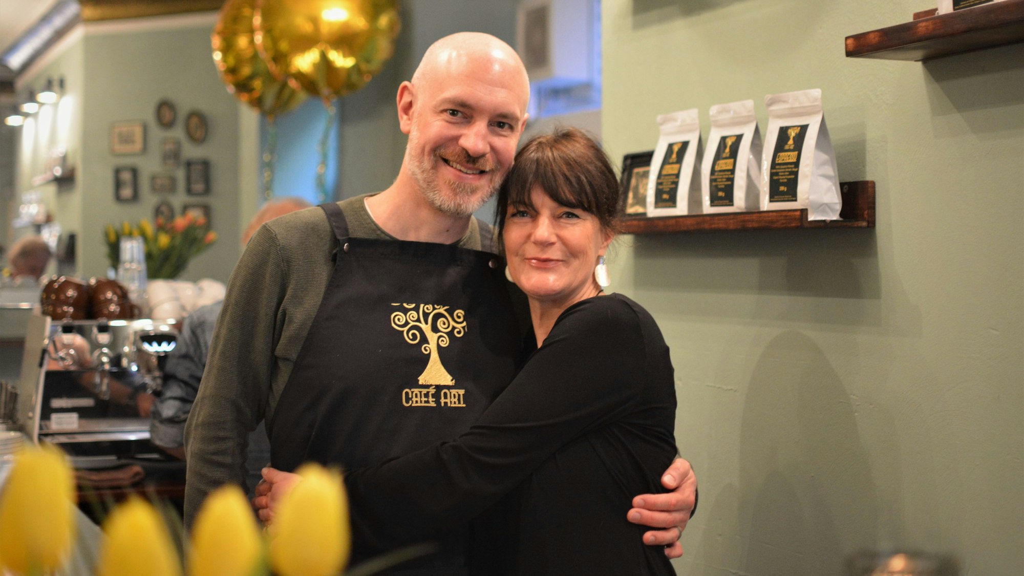 Das Ehepaar Kaiser/Klein umarmt sich in ihrem Café. Im Hintergrund sind Packungen ihres eigenen Kaffees zu sehen.
