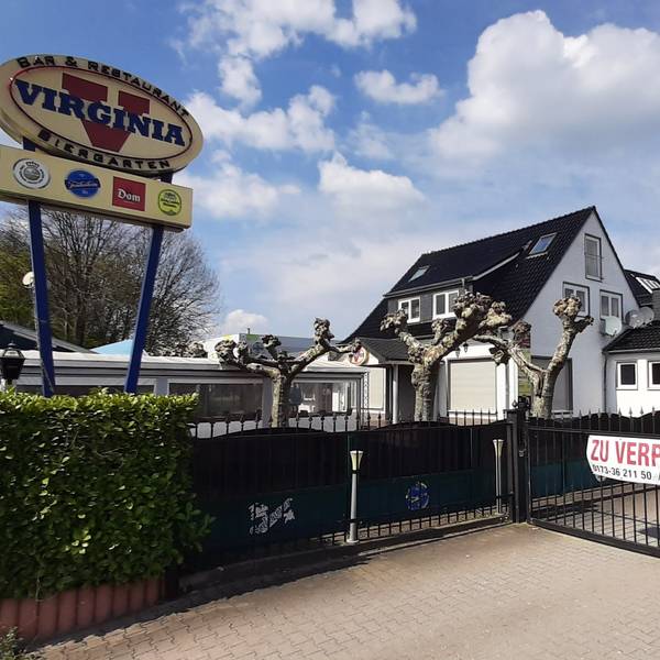 Das Virginia American Bar Restaurant in Monheim am Rhein befindet sich mittig hinter einem Zaun. Links findet sich ein Werbeschild mit dem Logo des Restaurants, rechts der Schotterparkplatz ohne Autos.