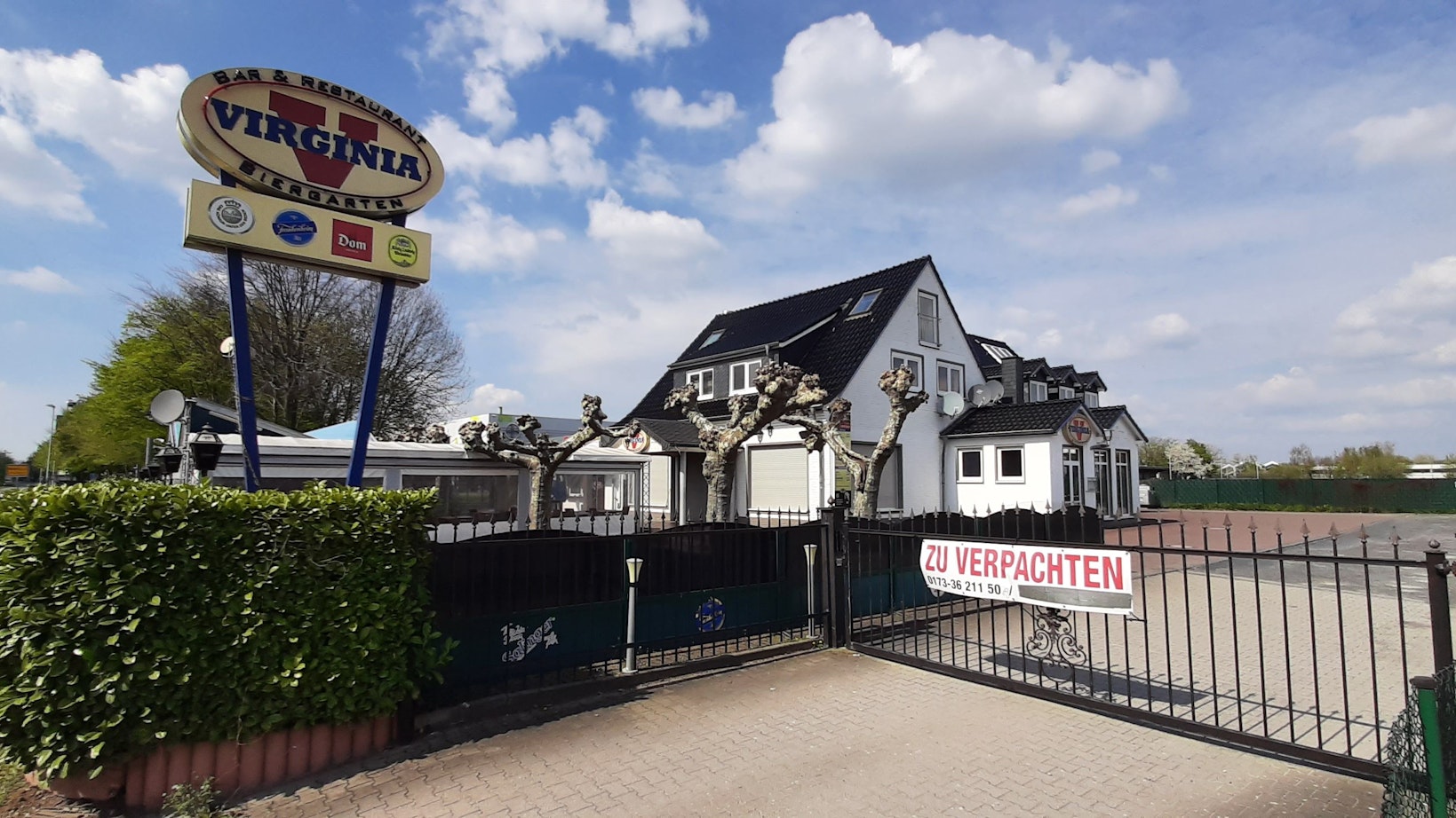 Das Virginia American Bar Restaurant in Monheim am Rhein befindet sich mittig hinter einem Zaun. Links findet sich ein Werbeschild mit dem Logo des Restaurants, rechts der Schotterparkplatz ohne Autos.